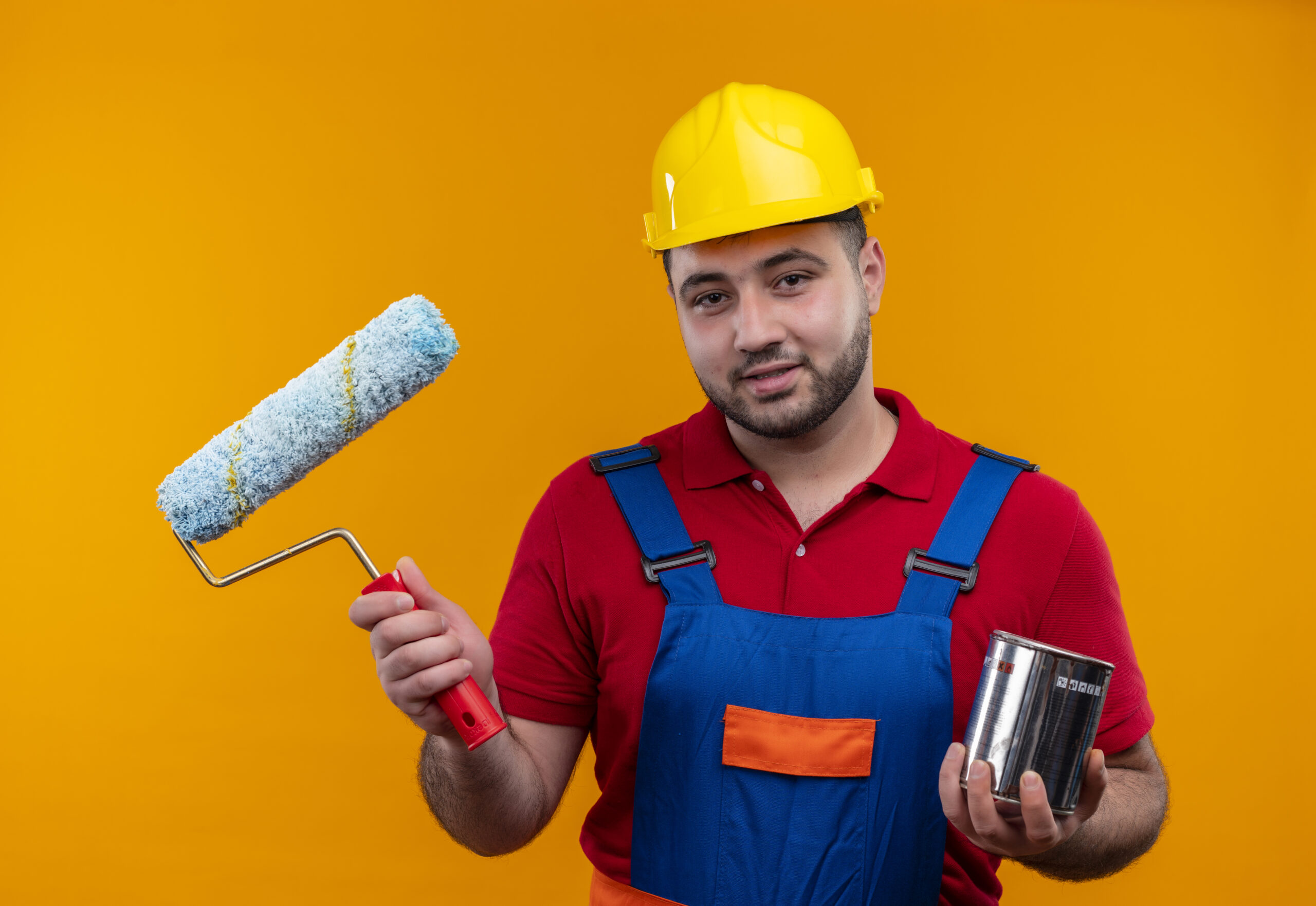 young builder man in construction uniform and safety helmetholding paint roller and paint can smiling standing over orange background