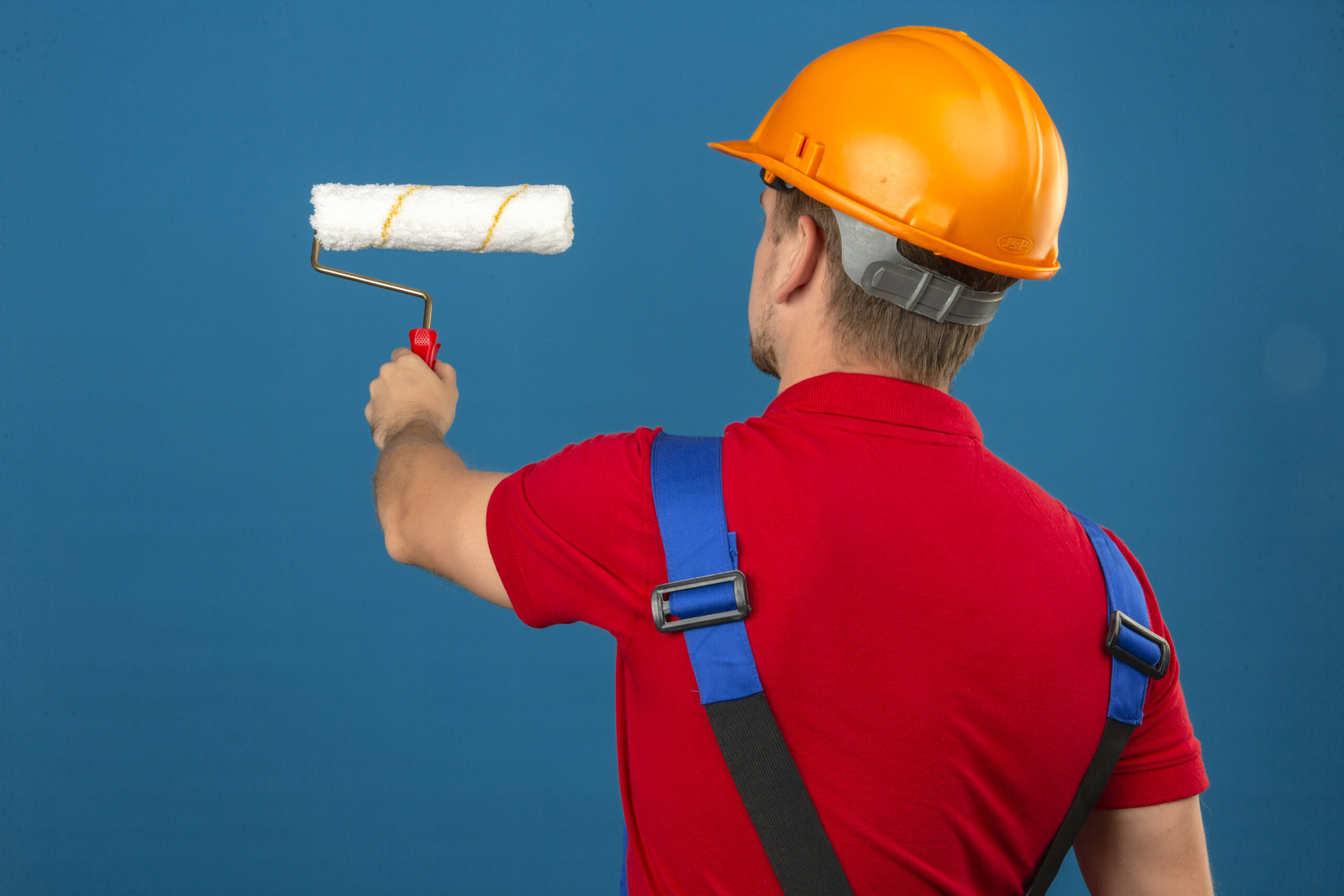 young builder man in construction uniform and safety helmet standing back with paint roller over isolated blue background